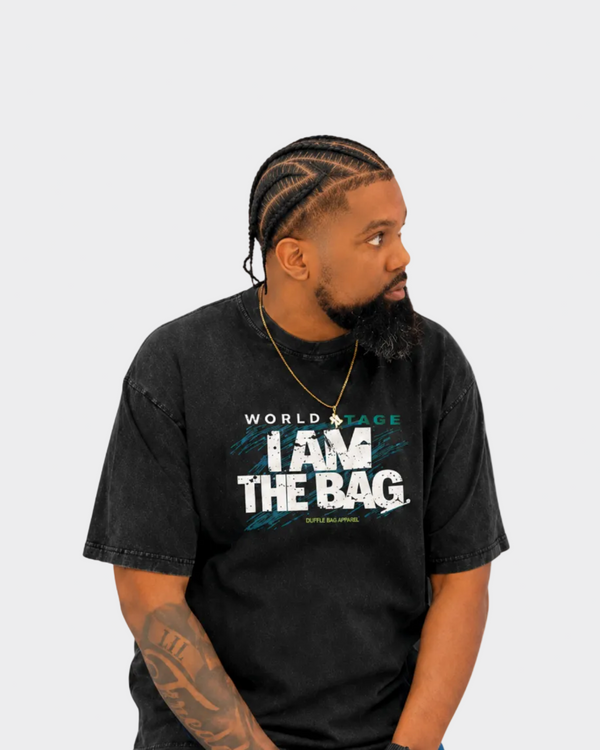 Man wearing a Duffle Bag black t-shirt with I AM THE BAG, sitting against a gray background